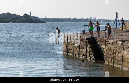 Musselburgh, East Lothian, Scozia, Regno Unito, 26 agosto 2021. UK Meteo: Giornata di sole per rinfrescarsi in mare. I bambini saltano fuori dalla parete del porto nel Firth of Forth durante il tempo caldo Foto Stock