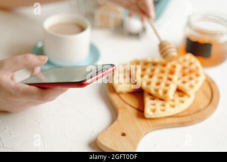 La donna scrive messaggi sul telefono delle cellule e la colazione. Mattina romantico passatempo. Comunicazione online a pranzo. Waffle belgi con miele, tazza di caffè su tavola bianca. Foto Stock