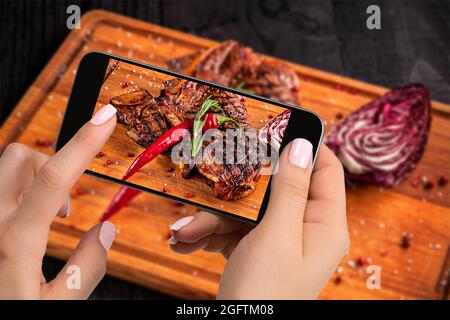 Fotografare il concetto alimentare - il turista scatta foto del piatto di bistecca di manzo pronto da mangiare sul taglio di legno a bordo di smartphone. Foto Stock