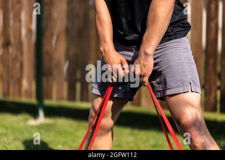 Primo piano del giovane uomo va in per lo sport a casa in cortile nel giorno d'estate. Giovane sportivo che fa squat con gomma sportiva su tappeto Foto Stock