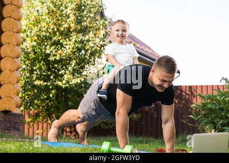 Il giovane uomo con bambino entra per praticare sport in giardino. Lo sportivo con capelli biondi si allena, facendo dei push-up con suo figlio sulla schiena sul prato Foto Stock