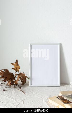Casa decoratore moccap, quadro vuoto cornice vicino bianco dipinto muro di cemento, rami con foglie asciutte e vecchi libri con cartoline Foto Stock