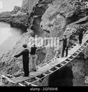 Una vista dei pescatori all'inizio del XX secolo e un cane che attraversa un ponte di corda più semplice a Carrick-a-Rede, vicino a Ballintoy nella contea di Antrim, Irlanda del Nord. Il ponte collega la terraferma alla piccola isola di Carrickarede, lunga 20 metri (66 piedi), si trova a 30 metri (98 piedi) sopra le rocce sottostanti. Foto Stock