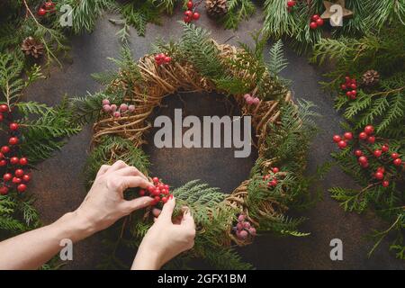 Donna fare la corona di Natale di fresco ramo di abete rosso naturale con bacche rosse su uno sfondo marrone. Anno nuovo. Vista dall'alto. Decorazione tradizionale festiva Foto Stock