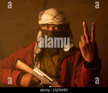 Felice militante o soldato islamico con la copertura facciale che mostra segno v o gesto di vittoria sulla guerra con la pistola in mano Foto Stock