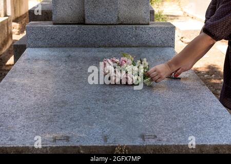 Le mani di una donna collocano un bouquet di fiori su una tomba di marmo in un cimitero. Si tratta di una tomba di marmo grigio con le iniziali RIP in latino e un out-of-foc Foto Stock