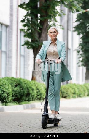 donna d'affari anziana in abito blu che guida scooter elettrico calcio all'aperto Foto Stock