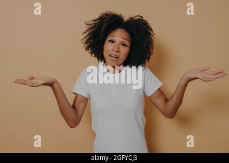 Incerto donna africana donna strugge spalle e stendendo braccia lateralmente, isolato su parete beige Foto Stock