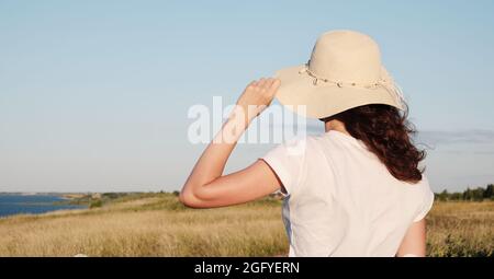 La ragazza si alza con la schiena che tiene il cappello e guarda il cielo al parapendio. Donna si alza con la schiena in un cappello su una collina. Foto Stock