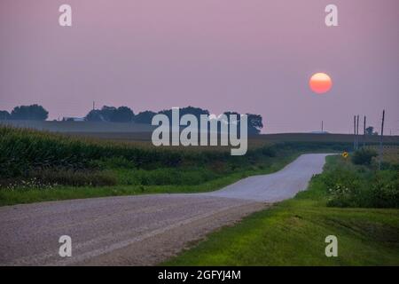 Il sole si imposta su una strada rurale in un cielo del Midwestern Iowa riempito di Haze dai fuochi della foresta della California, agosto 2021. Dyersville, Iowa, Stati Uniti. Foto Stock