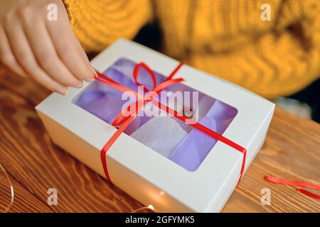 Ragazza in maglia arancione maglione apre regalo di festa. Natale o compleanno. Primo piano del nastro di estrazione manuale dalla scatola su tavola di legno. Foto Stock