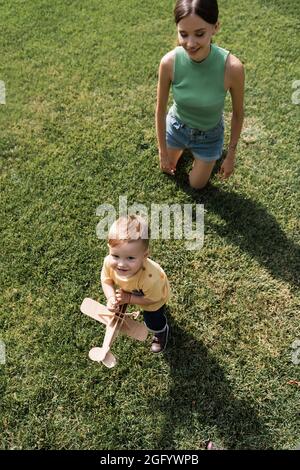 vista dall'alto della giovane madre felice che guarda il figlio allegro del bambino con biplano giocattolo e in piedi sull'erba Foto Stock