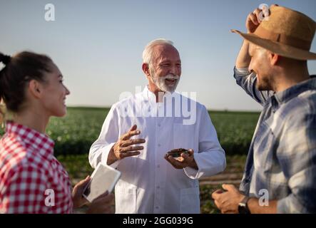 Donna agricoltore che utilizza una tavoletta in piedi sul campo. Giovane agricoltore maschio che tiene paglia cappello sorridente. Agronomo anziano che indossa un cappotto bianco con campione in Petri Foto Stock