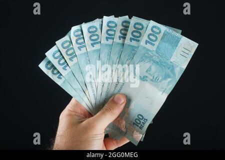 Mano che tiene soldi brasiliani. 100 banconote da reais. Sfondo nero. Foto Stock