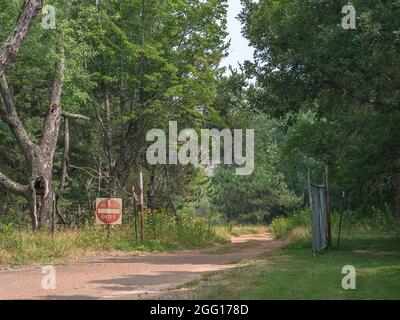 Il vialetto sporco vuoto e polveroso con il cartello "non entrare" e la recinzione conduce attraverso un cancello aperto in una foresta molto boscosa. Foto Stock