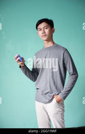ritratto di bell'uomo asiatico con t-shirt grigia con posa fredda. uomo skinny su sfondo verde. uomo con cellulare con espressione seria Foto Stock