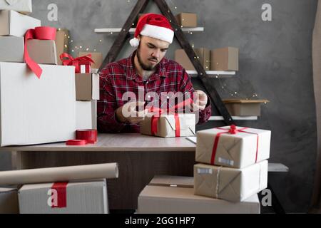 Un ragazzo in un cappello di Babbo Natale lavora a casa per imballare merci o regali per Natale per i bambini e lega un arco rosso con un nastro di seta su una scatola di cartone. S Foto Stock