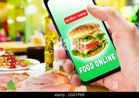 Tenere il telefono con l'app per ordinare e consegnare cibo su sfondo sfocato ristorante. Foto Stock