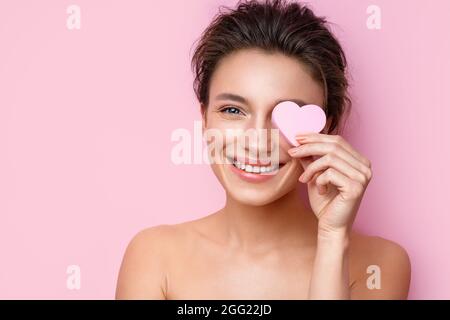 Donna sorridente con spugna cosmetica che copre un occhio. Foto di donna con perfetto trucco su sfondo rosa. Concetto di bellezza Foto Stock