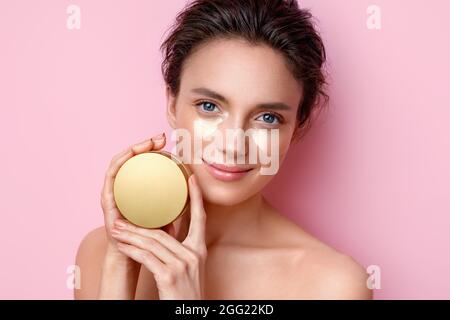 Donna con cerotti occhi che mostrano vaso con crema cosmetica. Foto di bella donna su sfondo rosa. Concetto di bellezza e cura della pelle Foto Stock