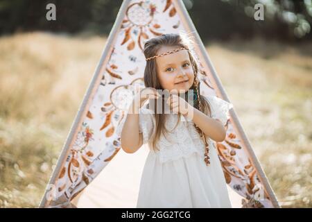 Primo piano scatto all'aperto di affascinante adorabile ragazza bambino, indossando abito bianco e piuma d'oca, in posa per la fotocamera vicino alla tenda da tè elegante in campo selvaggio estate bello Foto Stock