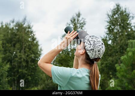 Giovane donna bionda uccello watcher in berretto e blu guardando attraverso binocoli al cielo nuvoloso in estate foresta ornitologica ricerca Birdwatching, zoog Foto Stock