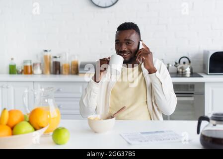 felice uomo afroamericano che tiene una tazza di caffè e parla sullo smartphone in cucina Foto Stock