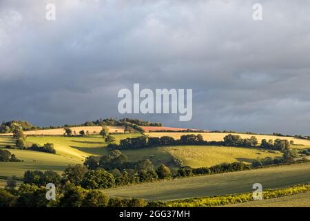 Una vista di terreni agricoli inglesi ondulati nella luce notturna di basso livello con un cielo grigio molto nuvoloso. Luce del sole sui campi. Foto Stock