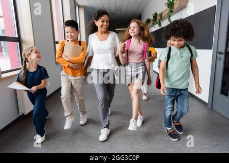 buon insegnante afroamericano che tiene le mani con la ragazza vicino gli allievi multietnici nella sala della scuola Foto Stock
