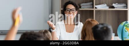 smiling african american teacher pointing with hand near blurred kids, banner Stock Photo