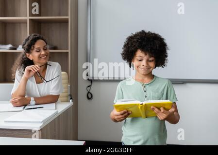 sorridente insegnante afroamericano che guarda il libro di lettura di schoolboy in aula Foto Stock