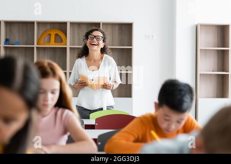 eccitato insegnante afroamericano che tiene il libro e ridendo durante la lezione Foto Stock