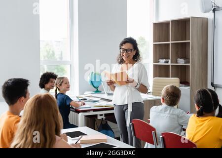 cheerful insegnante afroamericano che tiene il libro vicino ai capretti multietnici della scuola Foto Stock
