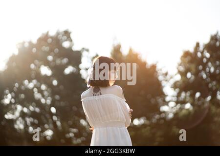 Ritratto posteriore della donna indiana americana nativa, in abito boho bianco, tatuaggi e capelli corti con piume, camminando in campo e foresta nel tramonto estivo. Natura, concetto di persone etniche Foto Stock