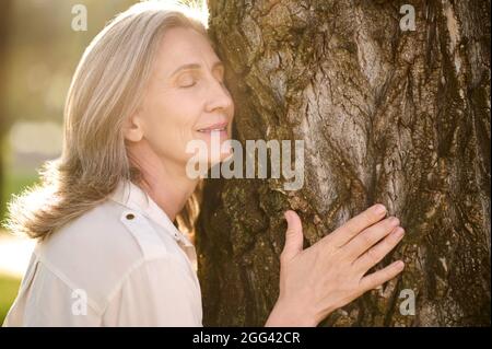 Donna sorridente adulta appoggiata all'albero Foto Stock