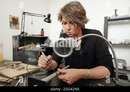 Gioielleria femminile concentrata seduta in officina con mobili neri e pezzi di saldatura insieme sotto lente d'ingrandimento Foto Stock