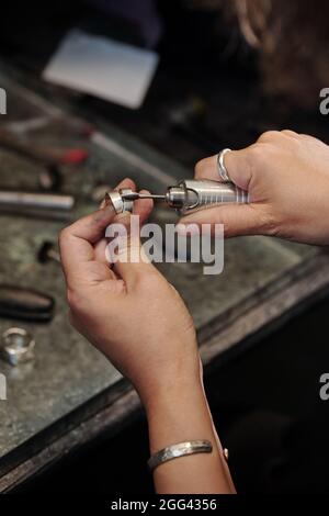 Primo piano di gioielliera femminile con mani sporche pulendo anello d'argento con lucidatrice in officina scura Foto Stock