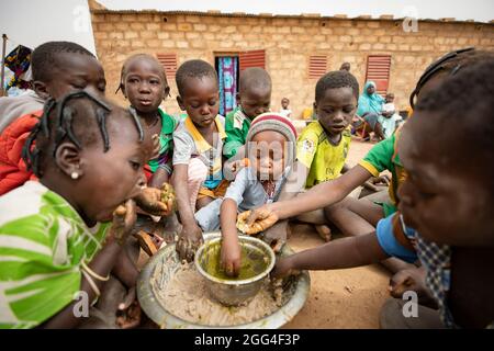 Un gruppo di bambini mangia intorno ad un piatto comune di pasta di miglio e stufato di foglie di baobab, probabilmente il loro unico pasto del giorno. Questa famiglia estesa è stata sfollata a causa della violenza e dell'insicurezza e ora si trova di fronte alla fame e alla carenza di cibo. Provincia di Kossi, Burkina Faso, Africa Occidentale. Foto Stock