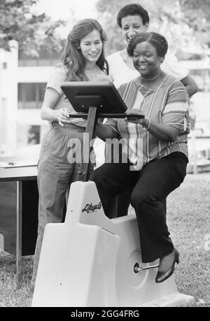 Austin Texas USA, circa 1990: La donna nera controlla la sua frequenza cardiaca mentre guida una bicicletta per esercizi Lifecycle ad una dimostrazione sponsorizzata dall'ospedale ad una fiera della salute in un parco cittadino. ©Bob Daemmrich Foto Stock