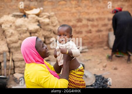 Una giovane madre che indossa un hijab islamico solleva sua figlia e sorride a lei fuori dalla loro casa a Nouna, Burkina Faso, Africa occidentale. Foto Stock