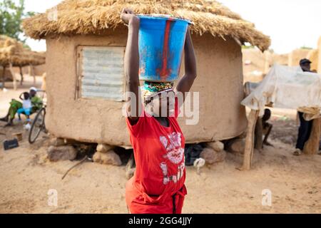 Mahammoud Traore (75, non raffigurato) sostiene una famiglia di 21 persone attraverso la sua agricoltura nel villaggio di Dougouninkoro, Mali. Ma il cambiamento climatico ha influenzato i modelli climatici negli ultimi anni e non è riuscito a crescere quanto prima. Ora, i negozi alimentari della sua famiglia si esaurono sempre prima di poter raccogliere il suo nuovo raccolto. Di conseguenza, ci sono sempre un paio di mesi ogni anno in cui la famiglia sperimenta la fame e spesso devono entrare in debito per comprare o prendere in prestito cibo. Qui, la figlia di Traore, Kadia (25), recupera l'acqua per la famiglia. Crisi Sahel 2021; Barouéli Cercle, Mali. Feb Foto Stock