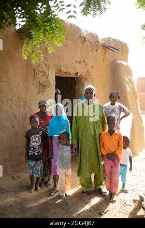 Mahammoud Traore (75, in verde) sostiene una famiglia di 21 persone attraverso la sua agricoltura nel villaggio di Dougouninkoro, Mali. Ma il cambiamento climatico ha influenzato i modelli climatici negli ultimi anni e non è riuscito a crescere quanto prima. Ora, i negozi alimentari della sua famiglia si esaurono sempre prima di poter raccogliere il suo nuovo raccolto. Di conseguenza, ci sono sempre un paio di mesi ogni anno in cui la famiglia sperimenta la fame e spesso devono entrare in debito per comprare o prendere in prestito cibo. Qui, il sig. Traore si alza con i membri della sua grande famiglia fuori della loro casa. Crisi Sahel 2021; Barouéli Cercle, Mali. Feb Foto Stock