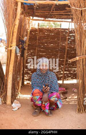 Il Senou Camp si trova al confine meridionale di Bamako, la capitale del Mali. Si tratta di uno degli otto campi informali della città e, di per sé, ospita 223 famiglie e poco più di 1.000 sfollati interni. Le famiglie che vivono qui sono fuggite dalla violenza e dall'insurrezione nella parte settentrionale e centrale del paese. Poiché la maggior parte di loro non ha mezzi di reddito o una normale fonte di cibo, la fame è una sfida quotidiana e la malnutrizione tra la sua popolazione continua ad essere diffusa. Qui, una donna siede nel suo rifugio di fortuna tenendo il suo telefono cellulare. Foto Stock