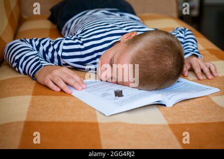 Il ragazzo si addormentò mentre si stese sul divano leggendo un libro o facendo i compiti. Il figlio si stancò dei suoi compiti e si addormentò. Bambino caucasico che è omeschooled Foto Stock