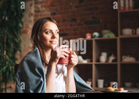 Giovane bella donna in sciarpa grigio accogliente con tazza di tè guarda fuori la finestra e riposa nel suo studio Foto Stock