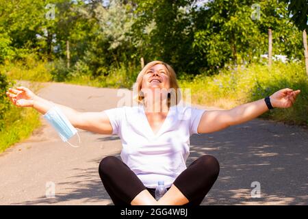 Donna anziana gioiosa che è nel parco seduto sulla strada che attraversa le gambe tenendo le mani sui lati godendo una giornata di sole con la maschera medica su uno Foto Stock