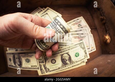 Mano che tiene cento banconote americane in rotolo sopra lo sfondo di cassetto di legno con lotto di dollari. Contanti di cento fatture del dollaro, valuta di soldi della carta. Foto Stock