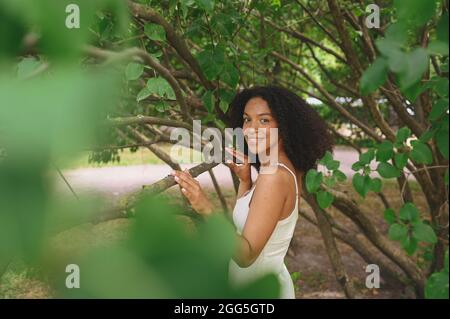 Moda primo piano ritratto di ridere attraente giovane naturalmente bella donna afroamericana con capelli afro e denti perfetti sorridono in natura parco in verde fogliame Foto Stock