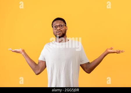 Confuso ragazzo nero che alza le mani e guarda la fotocamera, in piedi su sfondo giallo studio Foto Stock