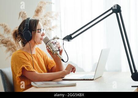 Podcaster femminile che registra il suo podcast utilizzando il microfono e il portatile presso il suo studio di trasmissione a casa. Donna in streaming dal vivo da casa studio radio Foto Stock